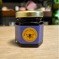 A small glass jar of bee products with a purple label and gold lid, sitting on a light wood tabletop with a blurred indoor background.