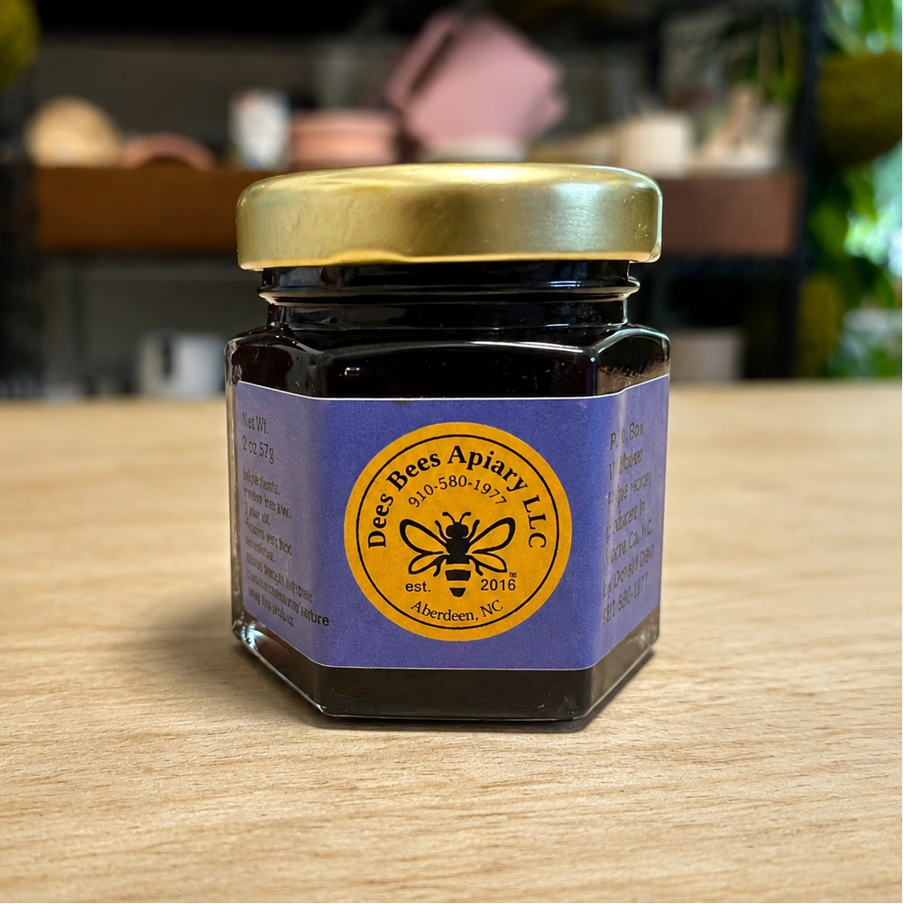 A small glass jar of bee products with a purple label and gold lid, sitting on a light wood tabletop with a blurred indoor background.